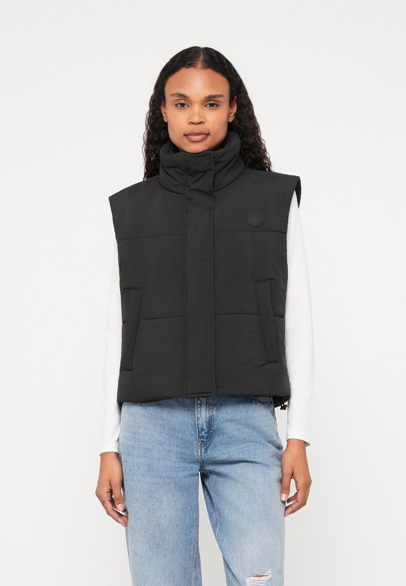 Μαύρο επενδυμένο γιλέκο με ψηλό γιακά, διαθέτει λογότυπο, δύο μπροστινές τσέπες και συντομευμένο σχέδιο. Φοριέται πάνω από ένα λευκό μπλουζάκι και τζιν παντελόνι.