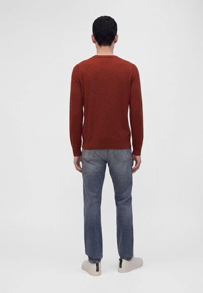 Homme debout, dos tourné, portant une chemise à manches longues couleur rouille, un jean bleu et des baskets blanches sur un fond clair uni.