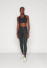 Top desportivo preto com design de costas nadador, combinado com leggings escuras estampadas com um tecido texturizado e uma cintura com o logótipo 'New Balance'. Ténis claros.