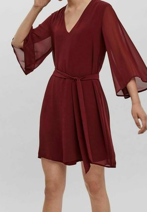 Kobieta ubrana w burgundową sukienkę do kolan z przezroczystymi, szerokimi rękawami do połowy przedramienia oraz pasem z tego samego materiału wiązanym w talii.