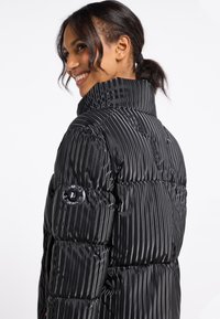 Czarna kurtka puchowa z pionowym wzorem, wysoki kołnierz i błyszcząca faktura. Posiada okrągłą łatę z logo na lewym rękawie.