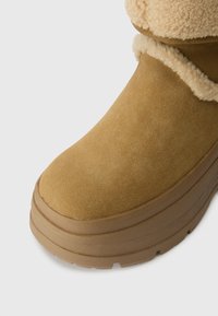 Bota de camurça em tom bege com um punho de pelúcia em creme. A bota possui uma sola grossa de plataforma com textura antiderrapante.