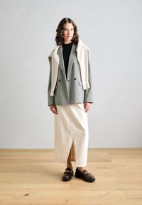 Gray houndstooth blazer over a black top, cream asymmetric skirt, and brown loafers with gold chain detail, set against a light background.