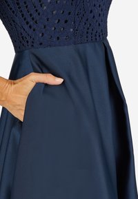Marineblaues Kleid mit einem strukturierten Häkeloberteil und einem glatten Satinstoffrock; die Hand in der Tasche zeigt eine funktionale Gestaltung mit verstecktem Detail.