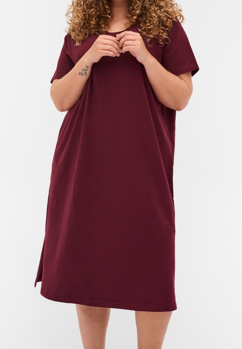 Femme aux cheveux bouclés portant une robe bordeaux ample, à manches courtes, longueur genou, mains jointes près de la poitrine, tatouage visible sur l'avant-bras.