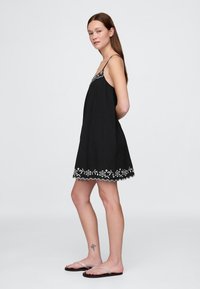 Črna obleka brez rokavov z vezenim belim cvetličnim vzorcem in valovito spodnjo robom. Lahka tkanina, tekoč dizajn in nastavljive naramnice.