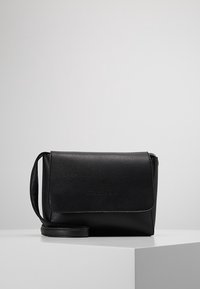 Czarna torebka crossbody z fakturowanej skóry, z klapką i regulowanym paskiem, wyeksponowana na białym cokole na szarym tle.