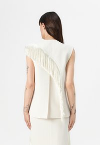 White fitted top with a diagonal fringe detail along the shoulder. Smooth texture, sleeveless design, and a clean back silhouette.