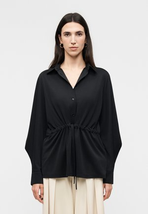 Γυναίκα που φοράει μαύρο πουκάμισο με κουμπιά και μακριά μανίκια, με ρυθμιζόμενο κορδόνι στη μέση, σε συνδυασμό με μπεζ πιετετέ παντελόνια με φαρδιά πόδια, μπροστά σε απλό φόντο.