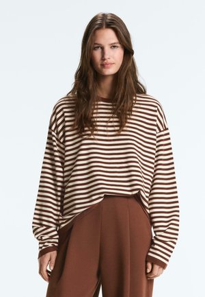 Giovane donna con lunghi capelli castani che indossa una maglia a maniche lunghe a righe marroni e bianche e pantaloni marroni a vita alta, rivolta verso il fronte.