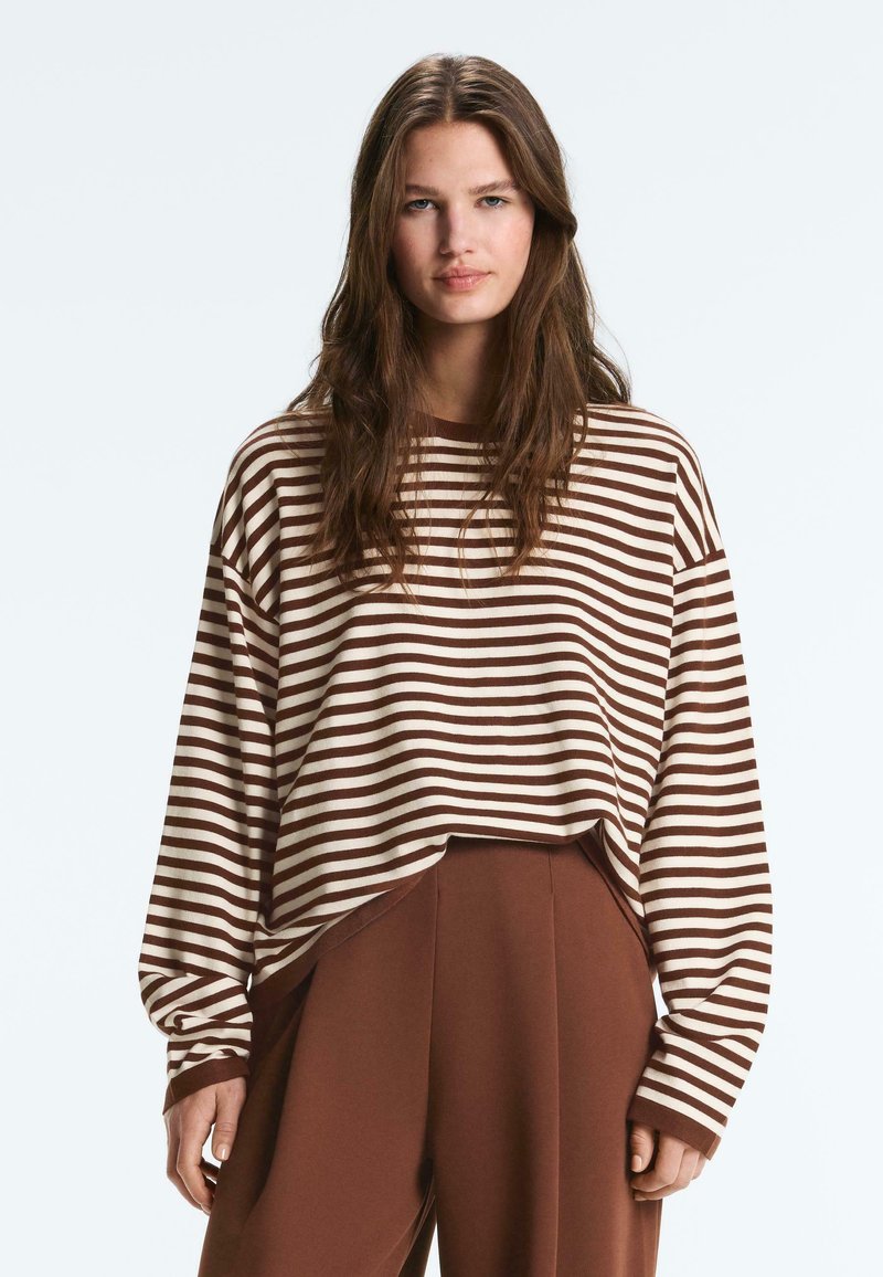 Fiatal nő hosszú barna hajjal, barna és fehér csíkos hosszú ujjú inget és barna, magas derekú nadrágot visel, előre néz.