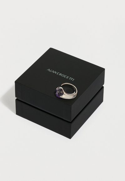 Ezüst gyűrű, amelyen egy hangsúlyos lila drágakő látható, fekete négyzet alakú dobozon van bemutatva, amelyen az ezüst színű márkanév olvasható.