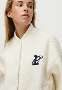 Valkoinen fleece-bomberitakki, jossa on ribbikaarroke, nappikiinnitys ja tummansininen brodeerattu logo vasemmassa rinnassa.