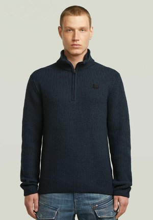 Man draagt een donkerblauwe, geribbelde gebreide trui met een kraag met halve ritssluiting en lichtblauwe denim cargobroek tegen een effen achtergrond.