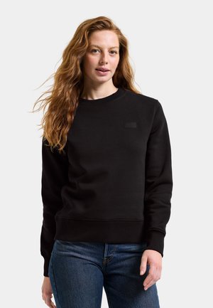 Kobieta o długich rudych włosach, ubrana w czarną bluzę z okrągłym dekoltem i niebieskie dżinsy, stojąca na tle jednolitego jasnego tła.