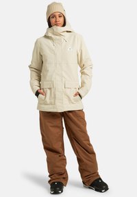 Kobieta w beżowej zimowej kurtce i czapce, w brązowych spodniach narciarskich i czarnych butach śniegowych, stoi z rękami w kieszeniach.