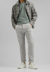 Grijze geruite jas over een groene henley-shirt, gecombineerd met lichtgrijze broek en witte sneakers. Recht gesneden, casual ontwerp met opgerolde mouwen.