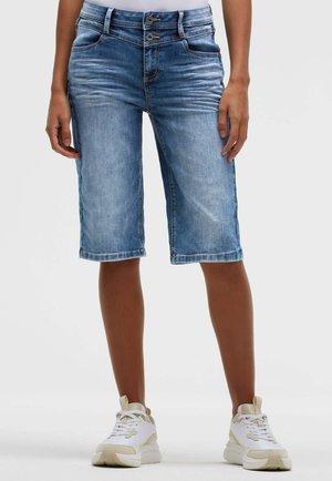 Person trägt blaue Denim-Shorts mittlerer Länge mit Doppelknopfverschluss an der Taille und weiße Sneakers, steht vor neutralem Hintergrund.