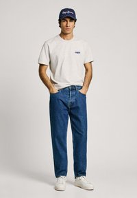 Light grey t-shirt with blue logo, paired with loose-fitting blue jeans and white sneakers. Model wearing a navy cap with white text.