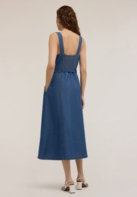 Robe en denim avec bretelles larges, buste ajusté avec smocks et jupe évasée, dans un bleu moyen. Poches sur les côtés et détails à l'arrière visibles.