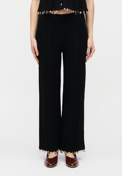 Γυναίκα που φοράει μαύρο πλεκτό παντελόνι καμπάνα με διακόσμηση από κοχύλια και χάντρες στο τελείωμα και ταιριαστό cropped τοπ, συνδυασμένο με καφέ παπούτσια.