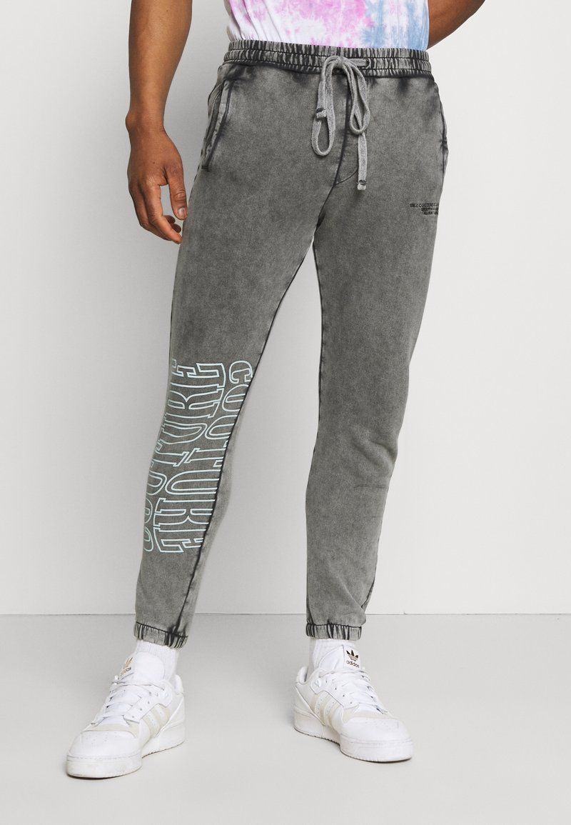 Šedé tepláky mají stahovací pas, elastické manžety a výrazný bílý textový design na jedné noze. Nosí se s bílými teniskami.