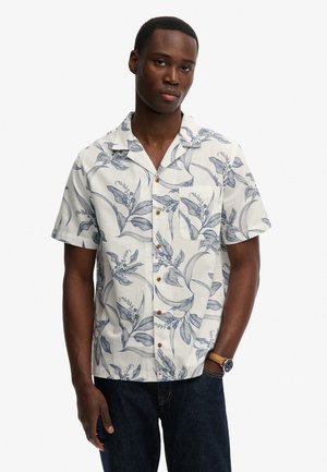 Man draagt een wit overhemd met korte mouwen met blauw botanisch motief en blauwe jeans, staat met één hand in de zak en een horloge om de pols.