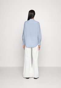 En person med långt mörkt hår står vänd bort, klädd i en ljusblå långärmad skjorta och vida vita byxor med svarta skor.