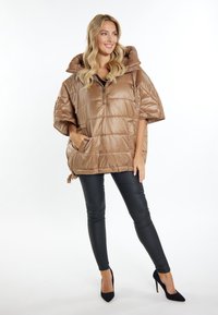 Gepolsterter, beige-brauner Poncho mit einem Reißverschlusskragen, übergroßer Kapuze und seitlichen Taschen. Kombiniert mit schwarzen, dehnbaren, eng anliegenden Hosen und schwarzen Absätzen.