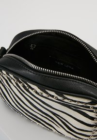 Černá taška z umělé kůže s přední stranou pokrytou kožešinou se zebra vzorem, ozdobená stříbrnými cvočky po obvodu. Obsahuje zapínání na zip.