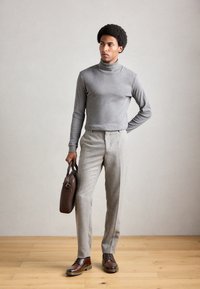 Hombre con suéter de cuello alto gris y pantalones beige sosteniendo un maletín de cuero marrón, de pie sobre un suelo de madera contra una pared gris claro.