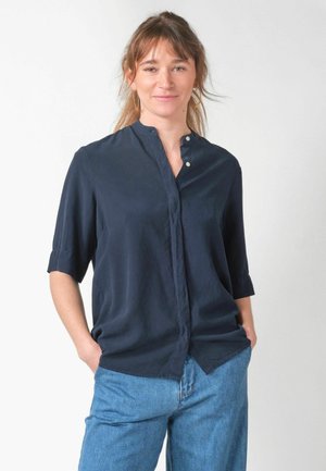 Kobieta o jasnobrązowych włosach, nosząca złote kolczyki koła, granatową koszulę z guzikami i rękawami do łokci oraz niebieskie dżinsy, stojąca z rękami w kieszeniach.