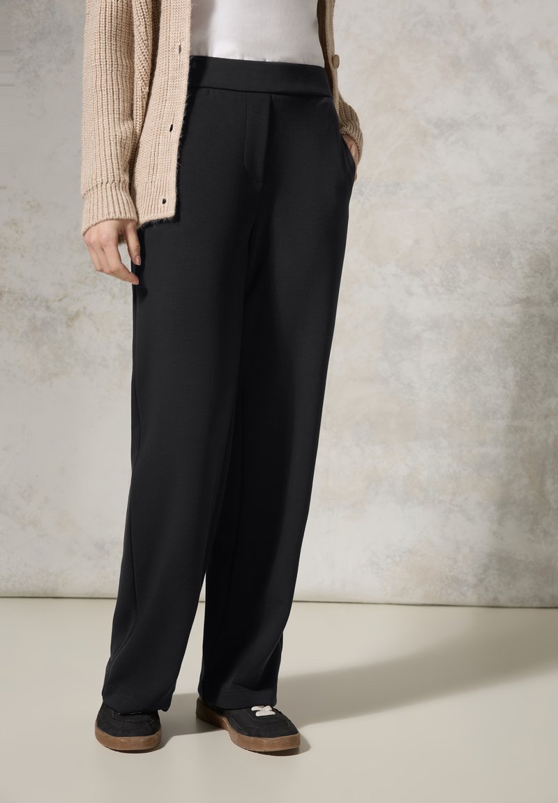 Pantalons larges noirs avec une texture lisse et des poches latérales. Associés à un cardigan en tricot beige et des chaussures noires avec un accent blanc.