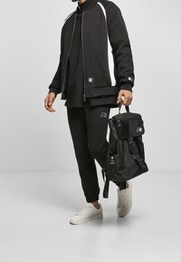 Chándal negro con detalles en blanco, combinado con una mochila negra. Calzado: zapatillas blancas. Tejido suave, ajuste holgado, diseño práctico.