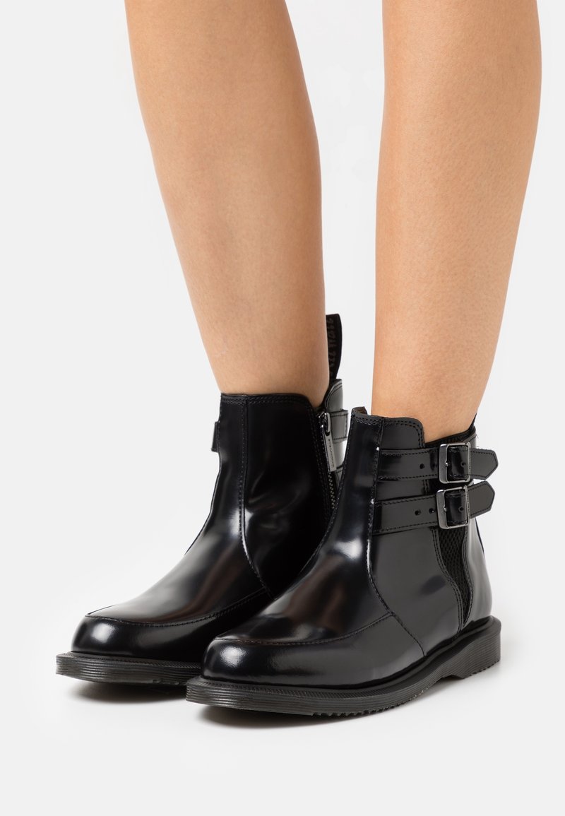 Bottes noires brillantes à chevilles avec fermetures éclair latérales et brides à double boucle portées sur des jambes nues sur fond blanc.