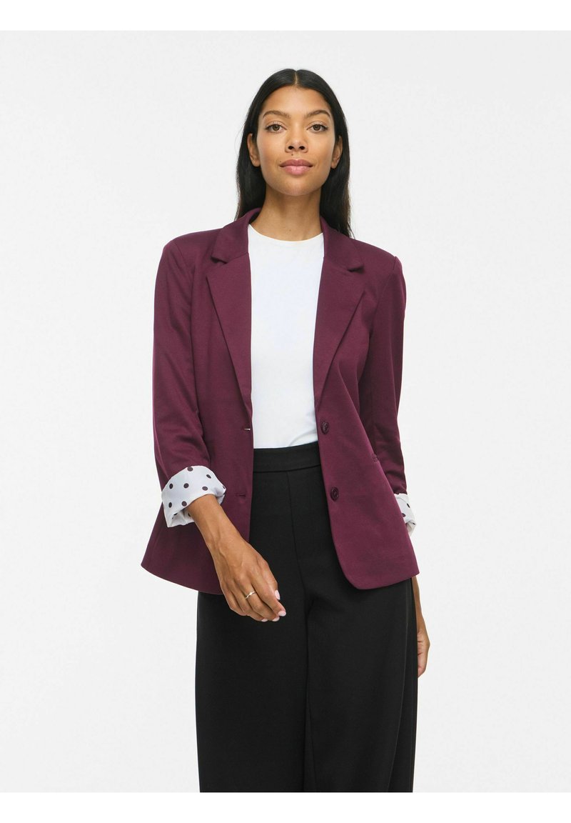 Femme portant un blazer bordeaux avec des poignets à pois, une chemise blanche et un pantalon noir, posant devant un fond blanc uni.