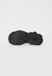 Musta kenkien pohja, jossa on teksturoitu pinta, kaarevia uria ja keskellä logo. Pääasiassa kumimateriaalia, joka on hieman joustavaa.