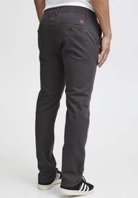 Gray chinos with a straight cut, featuring back pockets with button closures and a small brown logo patch. Paired with black and white sneakers.