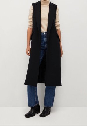 Personne portant un col roulé beige, un long gilet sans manches noir, un jean bleu et des bottines noires, debout devant un fond uni.