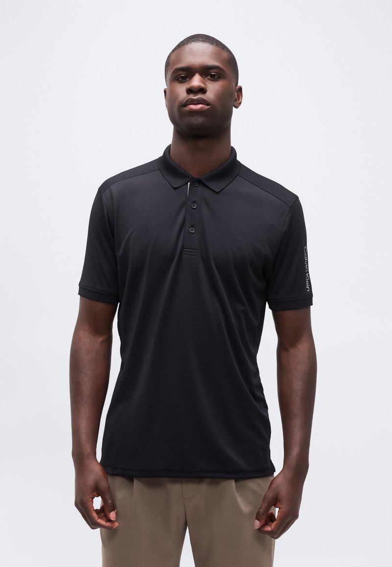 Άνδρας που στέκεται σε απλό φόντο, φοράει μαύρο κοντομάνικο πόλο και μπεζ παντελόνι, με το πρόσωπο προς τα εμπρός και τα χέρια χαλαρά δίπλα στο σώμα.