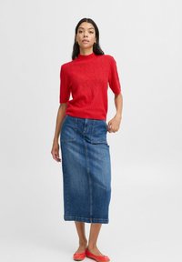 Rotes gestricktes Kurzarmoberteil mit strukturiertem Muster, kombiniert mit einem langen Jeansrock mit zwei Fronttaschen. Knallrote flache Schuhe.