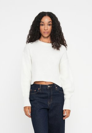 Hvit cropped genser med lange ermer, ribbestruktur og rund halslinning. Kombinert med mørkeblå denimjeans med kontrastsøm og metallknapper.
