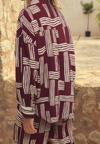 Chemise marron avec un motif de rayures abstraites blanches. Comprend un col, des manches longues et une coupe décontractée. Le tissu semble léger et texturé.