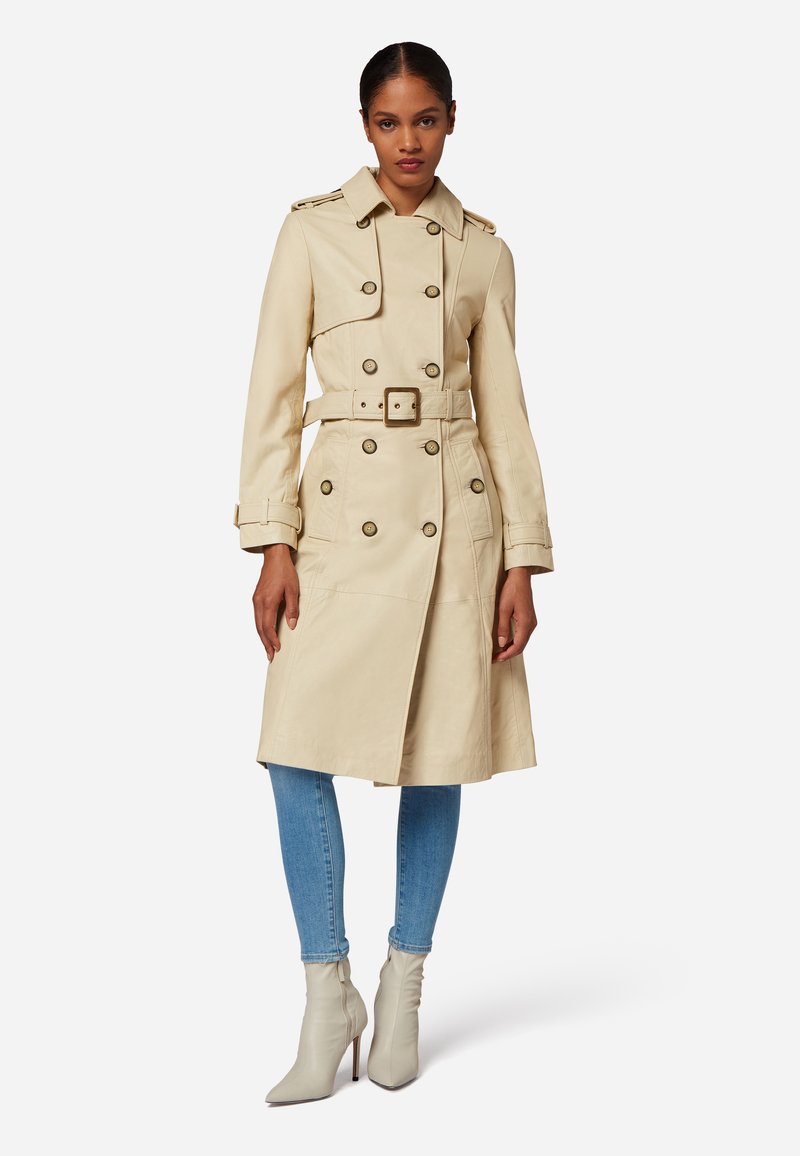 Beige trenchcoat med bälte i midjan, dubbelknäppt design, stora guldknappar och axelklapp, i kombination med ljusblå jeans och grå ankelstövlar.