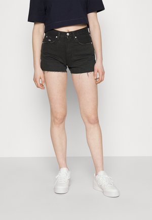 Pantalones cortos de mezclilla negra con dobladillo desflecado, bolsillos frontales y herrajes plateados. Combinados con zapatillas blancas. El modelo se presenta frente a un fondo liso.