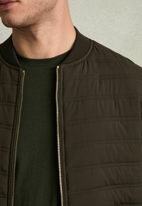 Jachetă bomber de culoare verde măsliniu închis, cu textură matlasată, guler ribat și fermoar complet. Pur și simplu peste un tricou verde asortat.