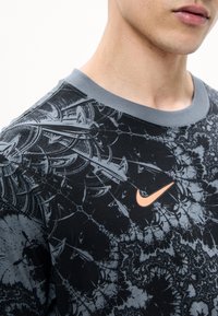 Κοντινό πλάνο ενός ατόμου που φοράει μαύρο και γκρι μπλουζάκι Nike με το λογότυπο Nike σε ροδακινί χρώμα κοντά στον γιακά.