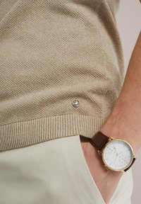 Bež pulover z pletenjem in ribanim robom, opremljen z majhnim srebrnim logotipom, v kombinaciji s svetlomi teksturiranimi hlačami in rjavim usnjenim uro.