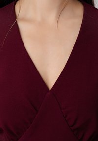 Haut bordeaux à col V avec un design croisé, arborant une texture lisse et des détails légèrement froncés au niveau de la poitrine.