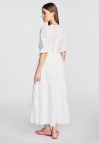 Biała bawełniana sukienka maxi z dziurkowanym wykończeniem, bufiastymi rękawami i falbanistą spódnicą. W zestawie z różowymi płaskimi butami, widok z tyłu.